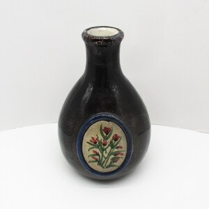 May include: A dark brown ceramic vase with a textured surface. The vase has a round body and a narrow neck. A circular design with a blue border features a floral pattern with red and green accents.