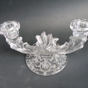 May include: Clear glass candle holder with two candle cups and a decorative floral base. The candle holder has a curved design with etched floral patterns. A classic home decor item.