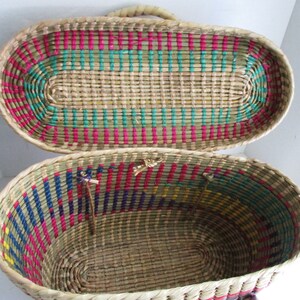 K&ouml;nnte beinhalten: Ein geflochtener ovaler Korb mit Deckel. Der Korb ist aus nat&uuml;rlichem Stroh mit bunten Streifen in Pink, Blau, Gr&uuml;n und Gelb. Der Deckel ist ge&ouml;ffnet und das Innere des Korbes ist sichtbar.