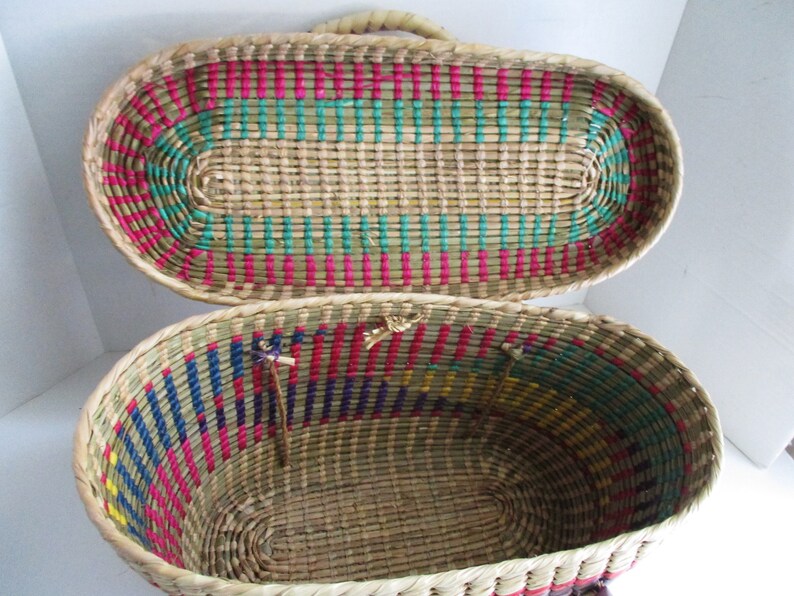K&ouml;nnte beinhalten: Ein geflochtener ovaler Korb mit Deckel. Der Korb ist aus nat&uuml;rlichem Stroh mit bunten Streifen in Pink, Blau, Gr&uuml;n und Gelb. Der Deckel ist ge&ouml;ffnet und das Innere des Korbes ist sichtbar.