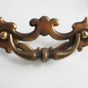 May include: A brass drawer pull with a decorative, ornate design. The pull has a curved handle and a center piece with a scroll design.