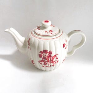 May include: White ceramic teapot with a lid, featuring a red floral pattern. The teapot has a curved spout and handle, and the body has a ribbed design. The lid has a small knob. A decorative item for tea lovers.