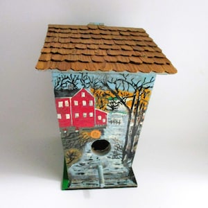 May include: Hand-painted wooden birdhouse with a brown shingled roof. The birdhouse features a scenic painting of a red building, trees, and a river. The birdhouse has a circular opening and a small perch.
