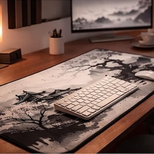Op de afbeelding: Een houten bureau met een grote muismat met een zwart-wit Japans landschapsontwerp. Een wit toetsenbord en muis liggen op de mat. Een lamp en een kleine plant zijn ook zichtbaar.
