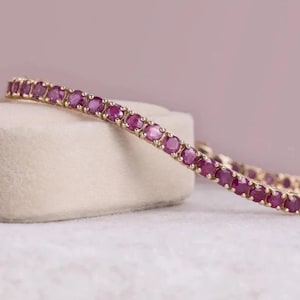 Puede incluir: Una pulsera de tenis que presenta una fila continua de piedras preciosas redondas de color rojo rosado intenso, engastadas en una banda de metal de oro amarillo. Las piedras son uniformes en tamaño y forma, sujetas por pequeñas garras.