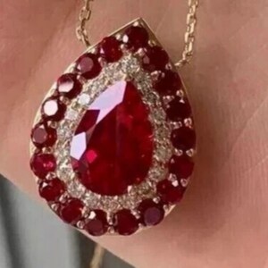 Puede incluir: Un colgante en forma de pera con una gran piedra preciosa roja central rodeada por un halo de pequeños diamantes blancos y un borde exterior de rubíes rojos redondos, montado en un marco de metal dorado con una fina cadena de oro.