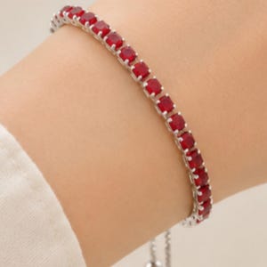 Könnte beinhalten: Ein silberfarbenes Tennisarmband mit einer durchgehenden Reihe quadratisch geschliffener, leuchtend roter Edelsteine. Das Armband verfügt über einen verstellbaren Schiebemechanismus mit zwei kleinen, runden silberfarbenen Perlen an den Enden.
