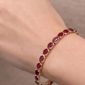 Puede incluir: Una pulsera de tenis en tono dorado que presenta una serie de piedras preciosas de color rojo intenso con corte ovalado, engastadas en una estructura de metal eslabonada. La pulsera tiene un diseño flexible con un patrón repetitivo de piedras rojas.