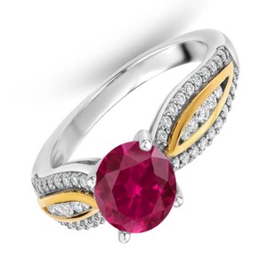 May include: A ring featuring a round, deep pink centre stone held by four prongs. The band is crafted from silver-toned metal with gold-toned accents shaped like leaves, and the shoulders are encrusted with small, clear, round-cut stones.