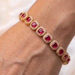 Puede incluir: Una pulsera de eslabones dorados con una serie de rubíes de color rojo intenso y corte cuadrado, cada uno rodeado por un halo de pequeñas piedras claras y brillantes. La pulsera está formada por segmentos metálicos articulados.