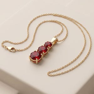 Puede incluir: Un collar de oro con un colgante de tres piedras redondas facetadas de color rojo intenso dispuestas en línea vertical. El colgante cuelga de una delicada cadena de oro que incluye un cierre de mosquetón para una sujeción segura.