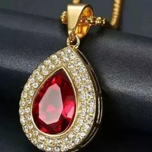 May include: A gold-toned pendant necklace featuring a large, pear-shaped, deep red faceted stone at the center. The stone is surrounded by a halo of small, clear, sparkling stones set in a gold-toned metal frame, suspended from a matching gold chain.