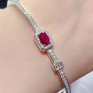 Może przedstawiać: Srebrzysta bransoletka typu bangle z centralnie umieszczonym, owalnie szlifowanym, ciemnoczerwonym rubinem otoczonym aureolą z małych, przezroczystych kamieni. Bransoletka jest ozdobiona dodatkowymi kamieniami oraz prostokątnym elementem z trzema kamieniami typu baguette.