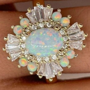 May include: A gold ring featuring a large central oval opal surrounded by a halo of small round opals and clear baguette-cut stones. The design includes additional small round clear stones interspersed between the opals and baguette-cut gems.