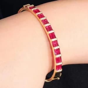 Puede incluir: Una pulsera rígida articulada de tono dorado que presenta una hilera de piedras rectangulares de color rojo vibrante engastadas en un canal. Cada piedra roja está separada por una fina barra dorada horizontal, creando un patrón.
