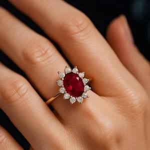 Könnte beinhalten: Ein Goldring mit einem großen, oval geschliffenen, tiefroten Rubin als Mittelstein, umgeben von einem Kranz aus zwölf kleinen, rund geschliffenen weißen Diamanten. Die Fassung besteht aus poliertem Gelbgold und bildet ein klassisches Blumenmuster.