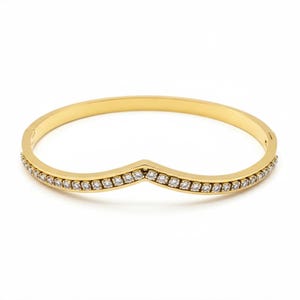 May include: A gold-toned hinged bangle bracelet featuring a V-shaped chevron design. The front section of the band is encrusted with a single row of small, clear, round-cut stones that follow the angular contour of the metal frame.