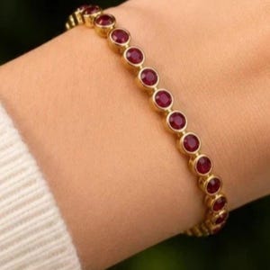 May include: A gold-toned bracelet featuring a continuous row of round, deep red gemstones set in individual circular bezels. The piece is designed with a flexible link structure, showcasing a uniform arrangement of stones along the entire length.