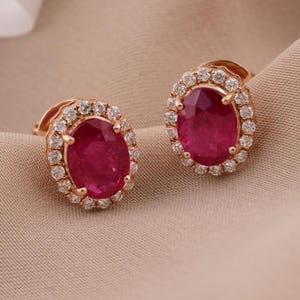 May include: A pair of oval-shaped stud earrings featuring deep red rubies set in rose gold. Each central ruby is surrounded by a halo of small, clear, round-cut diamonds, creating a classic and elegant design for these fine jewelry pieces.