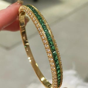 Op de afbeelding: Een gouden armband met een centrale rij vierkant geslepen groene stenen, geflankeerd door twee rijen kleine, ronde, heldere stenen. De metalen band heeft een gepolijste afwerking en een scharnierend ontwerp met een kanaalzetting.