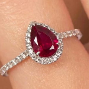 Pode incluir: Um anel com uma pedra central de rubi vermelho profundo em forma de pera, rodeada por um halo de pequenos diamantes brancos redondos. A banda é feita de metal prateado e também é cravejada com uma série de pequenos diamantes brancos no aro.
