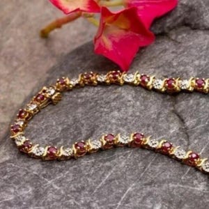 Op de afbeelding: Een gouden tennisarmband met een herhalend patroon van ronde rode robijnen en kleine heldere diamanten gezet in een metalen frame. De armband is in een halve cirkel gebogen en toont het afwisselende ontwerp van de edelstenen en schakels.