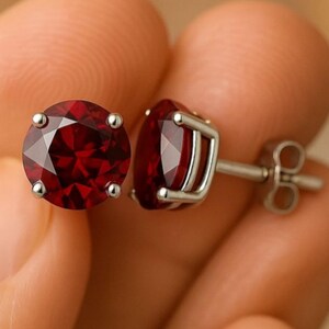 May include: A pair of round-cut deep red gemstone stud earrings set in silver-toned metal prongs. Each earring features a faceted stone held by four prongs, with a standard post and butterfly clutch backing visible on the side of one earring.