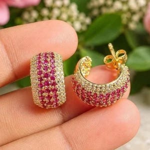 May include: A pair of gold hoop earrings featuring a wide band design. The surface of each hoop is encrusted with a central row of vibrant pink stones flanked by two outer rows of clear, sparkling stones, complete with a standard post backing.