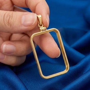 Puede incluir: Un marco de colgante rectangular en tono dorado con un borde interior liso y un borde exterior texturizado y acanalado. El colgante incluye una pequeña anilla articulada en la parte superior para sujetarlo a una cadena, con un acabado metálico pulido.