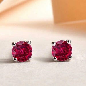 May include: A pair of round-cut, deep red gemstone stud earrings set in silver-toned metal. Each stone features a faceted surface and is secured by four small prongs, creating a classic circular design that highlights the vibrant crimson colour.