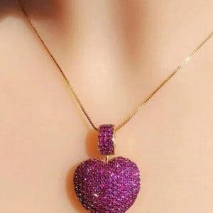 May include: A gold-toned chain necklace featuring a heart-shaped pendant. The pendant is fully encrusted with numerous small, vibrant magenta-colored stones, creating a textured, sparkling surface that covers both the heart and its bail.