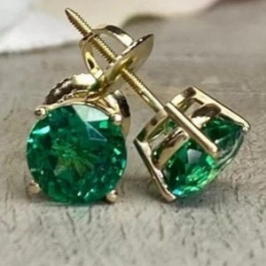 Peut inclure: Une paire de boucles d'oreilles à clous avec des pierres précieuses rondes, facettées et d'un vert éclatant, serties dans des paniers en métal doré. Chaque boucle possède une tige filetée et un fermoir à vis assorti en or.