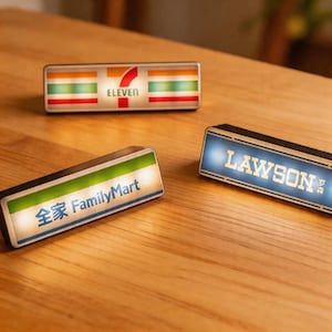 May include: Three illuminated rectangular signs featuring the logos of Japanese convenience stores: 7-Eleven, FamilyMart, and Lawson. The signs have white backgrounds, colourful borders, and text. The Lawson sign has blue backlighting.