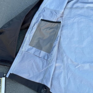 May include: Black and gray waterproof jacket with a hood. The jacket's interior is light gray with a pocket. The jacket is open, revealing the inside lining and construction. The jacket has a black zipper and a black trim.