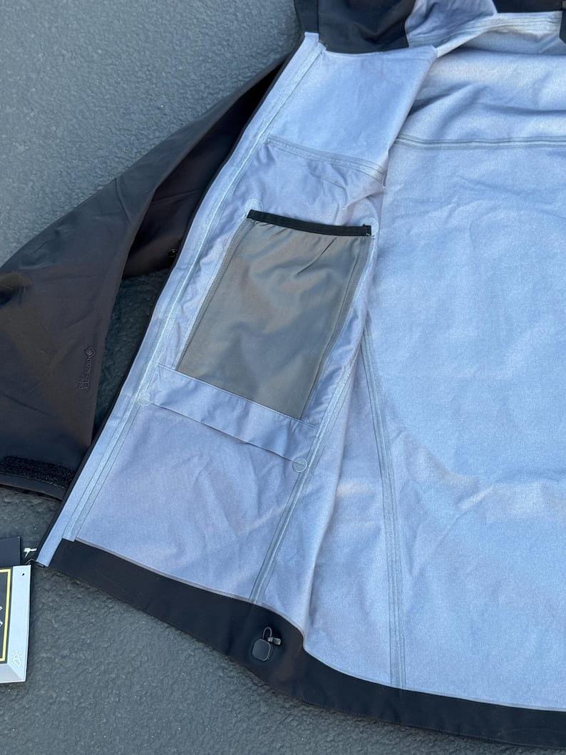 May include: Black and gray waterproof jacket with a hood. The jacket's interior is light gray with a pocket. The jacket is open, revealing the inside lining and construction. The jacket has a black zipper and a black trim.