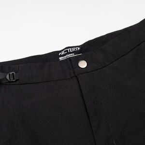 Pu&ograve; includere: Primo piano di pantaloni neri con chiusura a bottone e passante nero. I pantaloni hanno una tasca con cerniera e il marchio "Arc'teryx" &egrave; visibile sulla cintura. I pantaloni sono realizzati in un tessuto opaco e resistente.