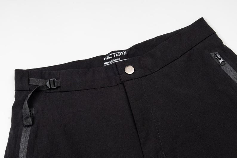 Pu&ograve; includere: Primo piano di pantaloni neri con chiusura a bottone e passante nero. I pantaloni hanno una tasca con cerniera e il marchio "Arc'teryx" &egrave; visibile sulla cintura. I pantaloni sono realizzati in un tessuto opaco e resistente.