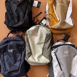 May include: Five backpacks in various colors, including black, navy blue, tan, and gray. Each backpack features a zippered main compartment, adjustable straps, and a logo on the front. The backpacks are displayed on a brown surface.