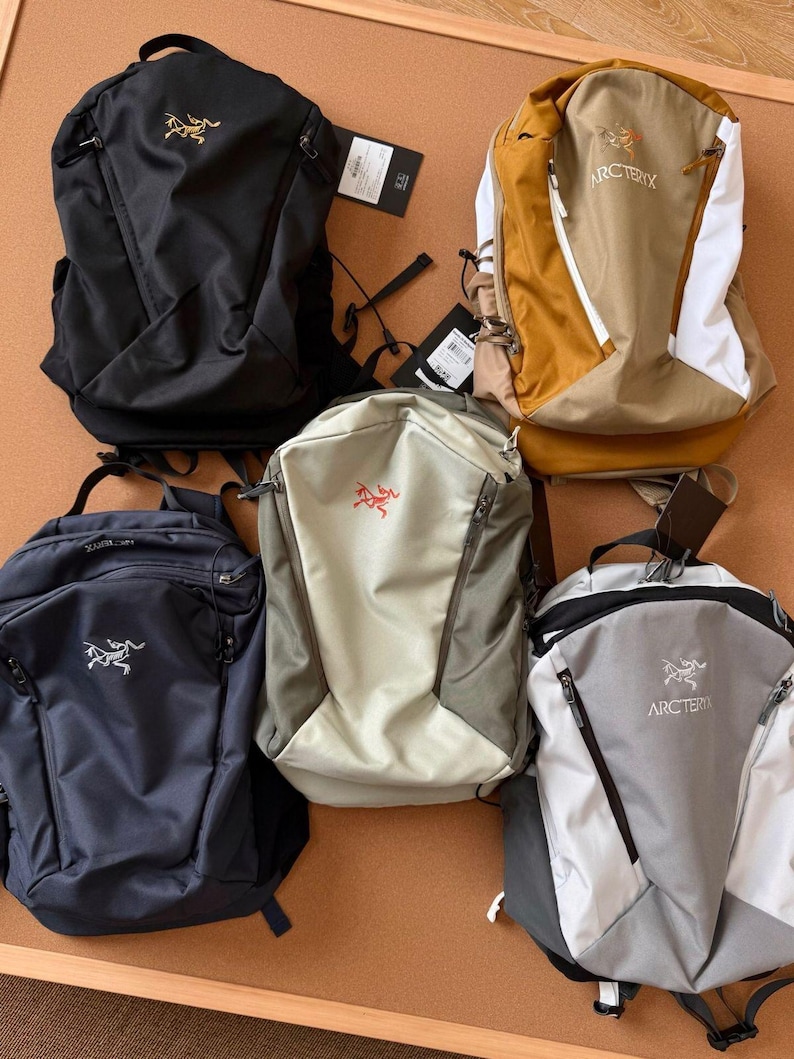 May include: Five backpacks in various colors, including black, navy blue, tan, and gray. Each backpack features a zippered main compartment, adjustable straps, and a logo on the front. The backpacks are displayed on a brown surface.