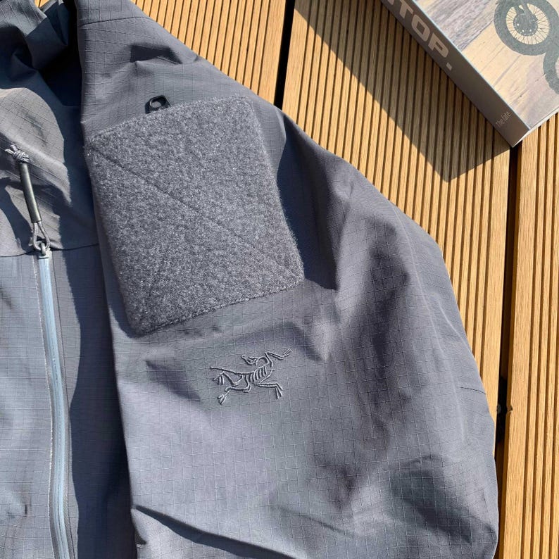 Op de afbeelding: Een grijze jas met een getextureerd oppervlak en een vierkante patch op de bovenarm. De jas heeft een ritssluiting en een klein logo op de mouw. De jas ligt op een houten oppervlak.