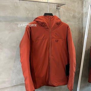 Op de afbeelding: Een roestkleurige jas met capuchon, een ritssluiting over de hele lengte en een klein logo op de borst. De jas hangt aan een metalen rek tegen een grijze muur met textuur. De merknaam "ARC'TERYX" is zichtbaar.