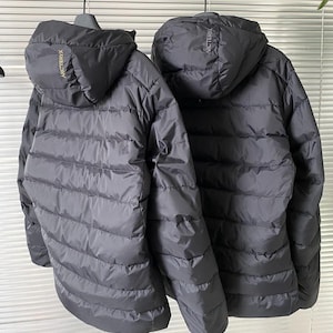Op de afbeelding: Twee zwarte pufferjacks met capuchon hangen naast elkaar. De jassen hebben horizontale gewatteerde stiksels en de merknaam "ARCTERYX" is in gouden letters op de capuchon gedrukt. De jassen zijn ontworpen voor koud weer.