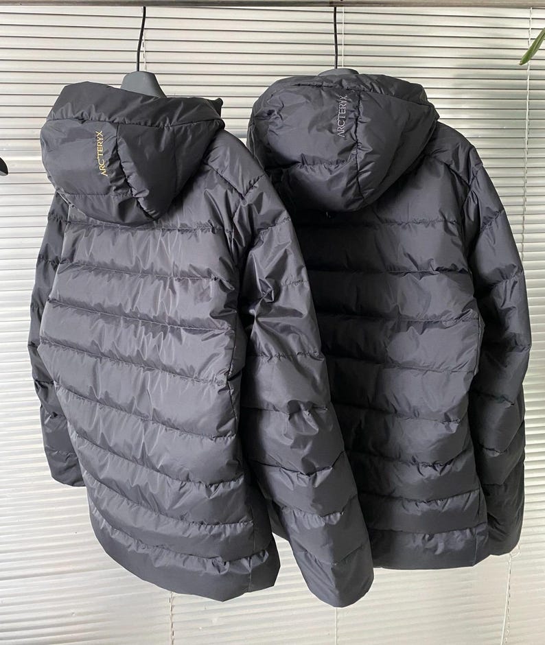 Op de afbeelding: Twee zwarte pufferjacks met capuchon hangen naast elkaar. De jassen hebben horizontale gewatteerde stiksels en de merknaam "ARCTERYX" is in gouden letters op de capuchon gedrukt. De jassen zijn ontworpen voor koud weer.