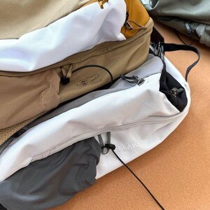 May include: A close-up of several backpacks in various colors, including tan, white, and gray. One backpack has the word "ARC" printed on it, and another has the word "BEAMS" printed on it. The backpacks have zippers and drawstrings.