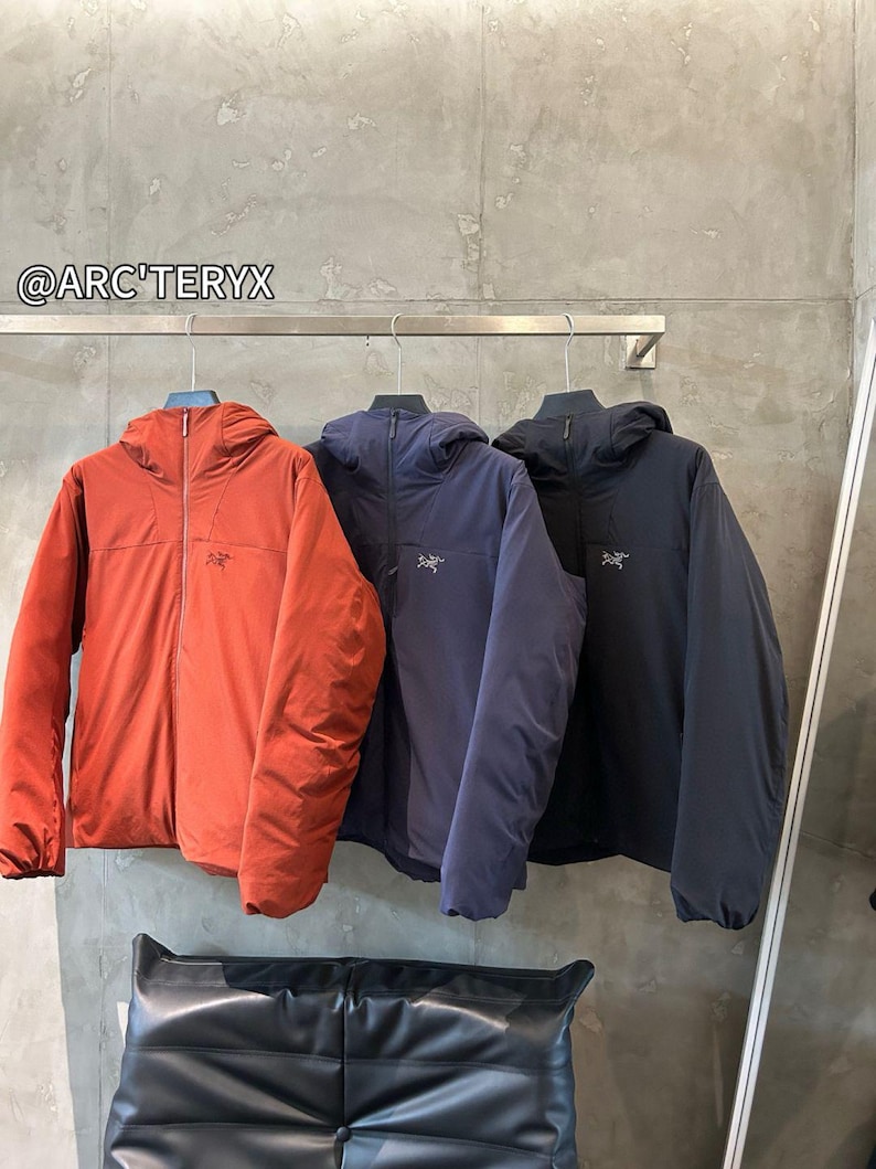 Op de afbeelding: Drie jassen met capuchon in roest, marineblauw en zwart hangen aan een metalen stang. Elke jas heeft een volledige ritssluiting en een klein logo op de borst. De jassen worden getoond tegen een neutrale grijze muur. De merknaam "@ARC'TERYX" is zichtbaar.