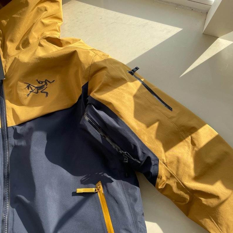 May include: A navy blue and mustard yellow jacket with a hood and multiple zipped pockets. The Arc'teryx logo is visible on the chest. The jacket is designed to be waterproof and is suitable for outdoor activities. The jacket has a modern design.
