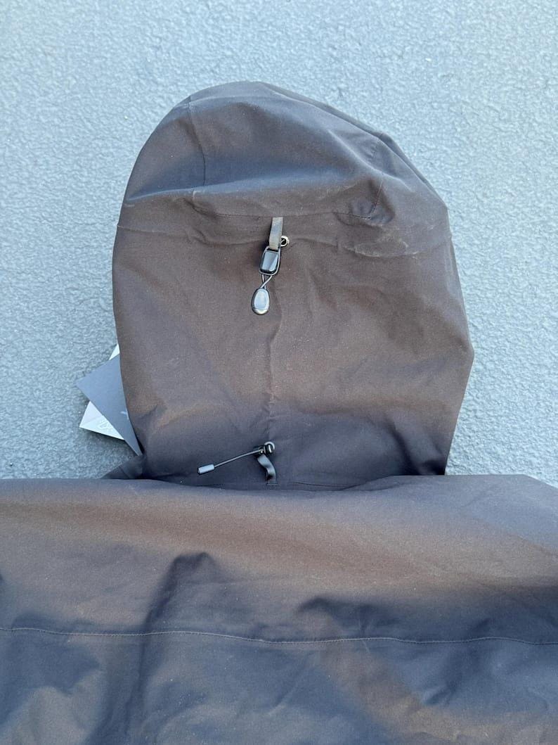 May include: Close-up of a black hooded jacket. The hood is up, revealing a drawstring and a small, decorative toggle. The jacket appears to be made of a durable, water-resistant material. The hood is attached to the jacket with a seam.