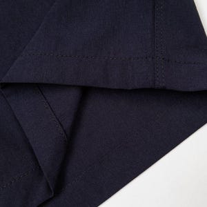 Pu&ograve; includere: Primo piano di un tessuto blu navy, che mostra l'orlo piegato e le cuciture. Il tessuto ha una trama sottile e le cuciture sono visibili lungo i bordi, evidenziando la costruzione del capo.