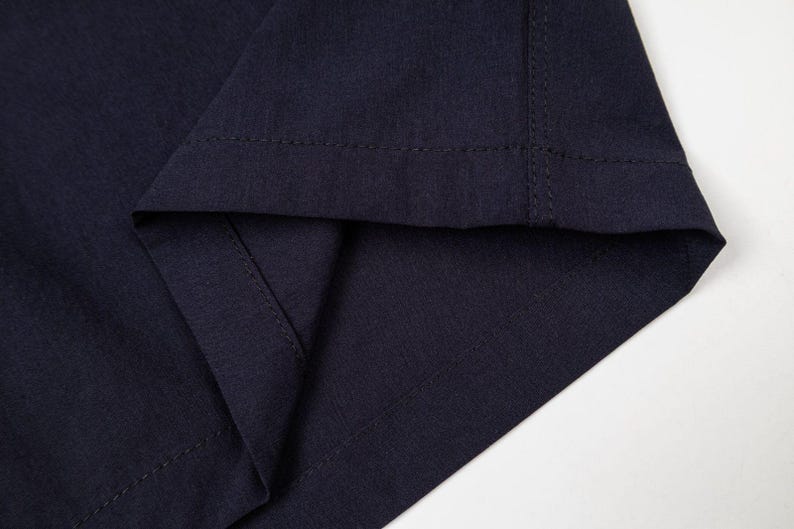 Pu&ograve; includere: Primo piano di un tessuto blu navy, che mostra l'orlo piegato e le cuciture. Il tessuto ha una trama sottile e le cuciture sono visibili lungo i bordi, evidenziando la costruzione del capo.