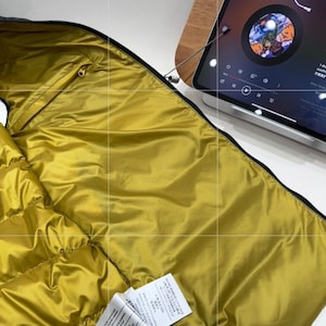 May include: Close-up of a mustard-yellow puffer jacket interior with black trim and a gold zip. The jacket is partially open, showing the quilted lining. Two white tags are visible.